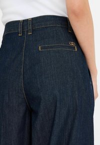 Pantaloni in denim blu scuro a gamba dritta, con cuciture gialle, tasca posteriore e un piccolo logo embossato sul cinturino.