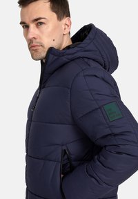 Giacca imbottita blu navy con cappuccio, caratterizzata da una texture trapuntata, tasche con zip e una toppa verde con la scritta "URBAN WEAR" sulla manica.