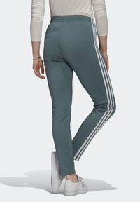 Pantaloni jogger in teal con una vestibilità slim, cintura elastica e tre strisce bianche laterali, abbinati a scarpe da ginnastica bianche. Tessuto liscio e leggero.