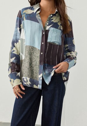Woman wearing a multicolored patchwork button-up shirt with floral and striped patterns, paired with dark blue wide-leg jeans.
