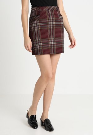 Mini-jupe écossaise rouge à carreaux avec poches, présentant des lignes verticales et horizontales en blanc, jaune et accents bleus. Associée à des chaussures à talons noires.