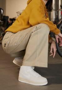 Białe wysokie trampki, beżowe bawełniane spodnie i żółta bluza z kapturem. Skoncentruj się na fakturze i kolorach, z sznurówkami i kontrastującymi detalami przeszyć.