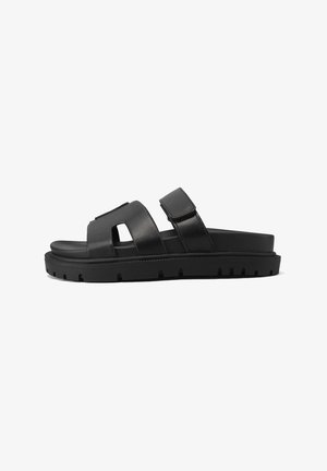 Sandali in pelle nera con una larga fascia caratterizzata da un design a intagli e chiusura regolabile, montati su una suola in gomma testurizzata.