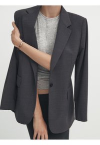 Blazer grigio scuro con colletto a tromba, chiusura a un bottone e tasca frontale. Indossato sopra una maglietta grigia, che mette in mostra una vestibilità su misura.