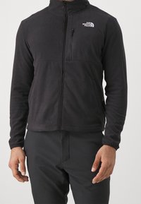 Mężczyzna ubrany w czarną polarową kurtkę z zamkiem błyskawicznym marki North Face, z kieszenią na piersi i czarne spodnie, stoi na jednolitym tle.