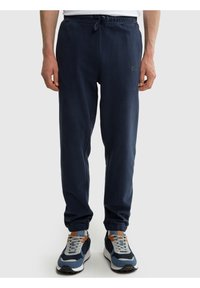 Vyras vilki tamsiai mėlynas sportines kelnes su elastinėmis rankogaliais ir raišteliu juosmens srityje, dėvi mėlynas ir pilkas sportines basutes, stovi priešais baltą foną.