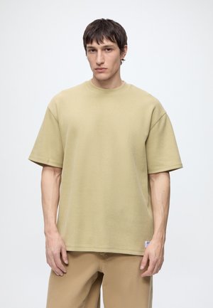 Giovane uomo con capelli corti e scuri che indossa una camicia beige a maniche corte e pantaloni abbinati, in piedi davanti a uno sfondo chiaro e uniforme.