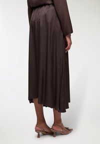 Abito marrone, alla lunghezza del ginocchio, con orlo fluido, realizzato in tessuto morbido. Presenta delle pieghe sul retro ed è abbinato a tacchi beige chiari.