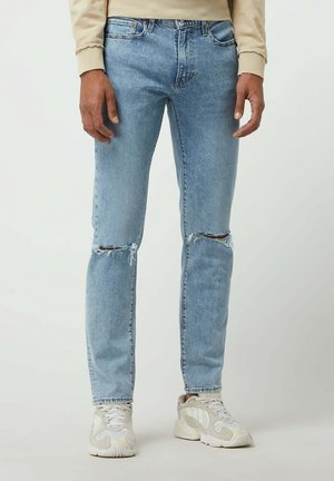 Homme portant un jean déchiré bleu clair et des baskets beiges, debout devant un fond clair uni.