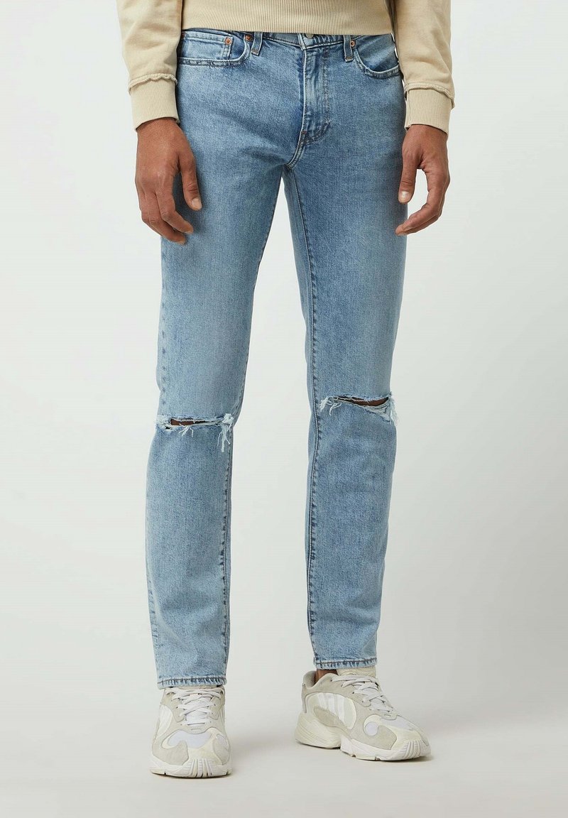 Homme portant un jean déchiré bleu clair et des baskets beiges, debout devant un fond clair uni.