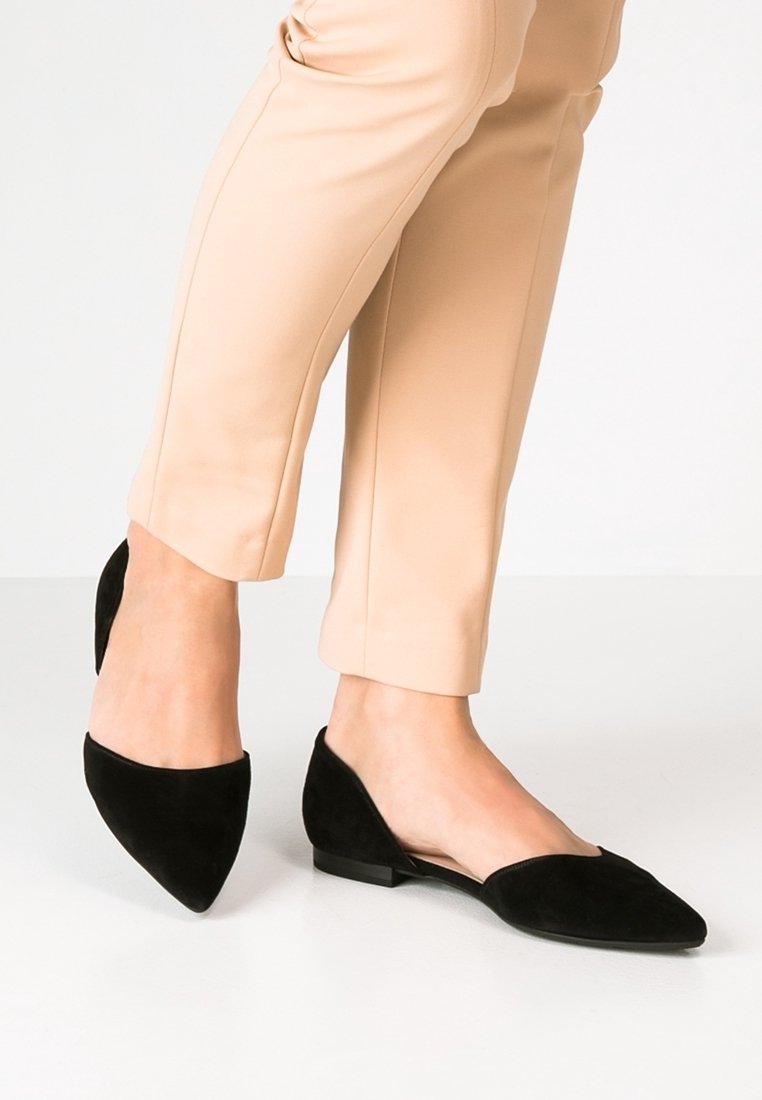 Pieds portant des chaussures plates noires à bout pointu, associées à un pantalon beige longueur cheville, sur un fond blanc.