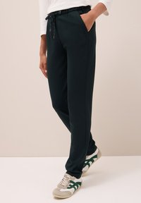 Tmavě zelené tepláky vyrobené z hladké látky, s elastickým pasem na šňůrku, bočními kapsami a zúženým střihem nohavic.