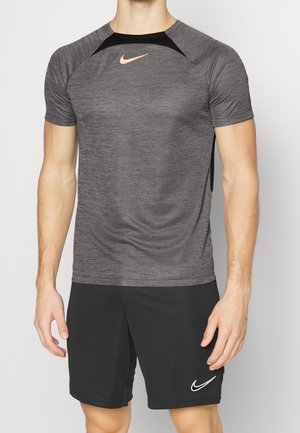 Mężczyzna w szarej sportowej koszulce Nike i czarnych szortach Nike stoi na tle jednolitego jasnego tła.