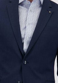Veste de costume bleu marine sur une chemise blanche à boutons avec un motif paisley bleu clair, boutonnée au col et à l'avant de la veste.