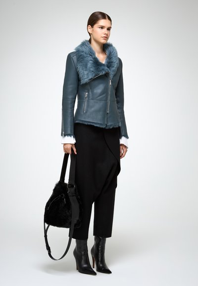 Giacca di pelle teal con colletto in pelliccia sintetica, tasche con zipper, abbinata a pantaloni neri testurizzati e stivali neri con tacco alto. Borsa nera.