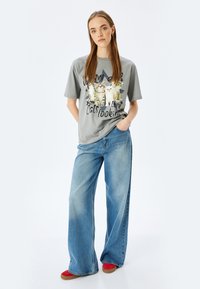 Camiseta gráfica gris con gatos, combinada con unos pantalones vaqueros de corte holgado de color azul claro y zapatos rojos, de pie frente a un fondo blanco.