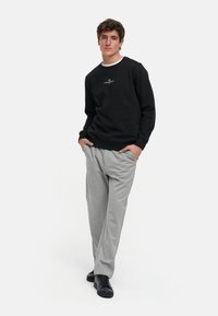 Sudadera negra de cuello redondo, pantalones de chándal grises de corte relajado y zapatillas negras, llevadas por una persona de pie con las manos en los bolsillos.