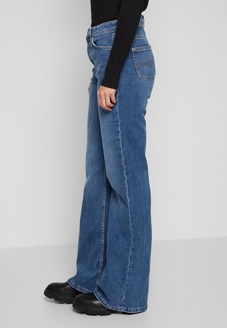 Jean en denim bleu taille haute avec une coupe flare, comportant cinq poches et une texture lisse. Des chaussures noires complètent le look.