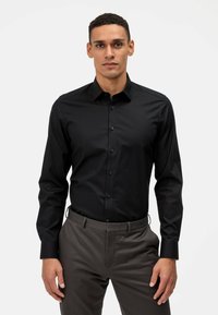 Chemise noire à manches longues avec une coupe ajustée, fermeture à boutons sur le devant et une texture lisse, associée à un pantalon gris, créant un look élégant.