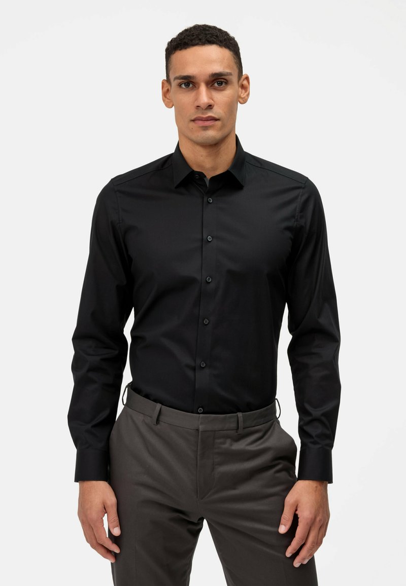 Chemise noire à manches longues avec une coupe ajustée, fermeture à boutons sur le devant et une texture lisse, associée à un pantalon gris, créant un look élégant.