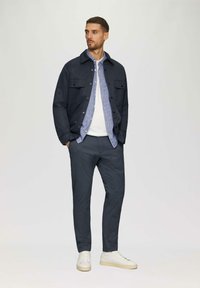 Mariene katoenen jas over een lichtblauw geruit shirt, wit t-shirt met ronde hals en donkere broek, gecombineerd met witte sneakers.