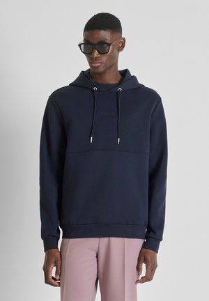 Uomo con occhiali da sole neri, felpa blu navy con coulisse e pantaloni rosa chiaro, in piedi contro uno sfondo chiaro uniforme.