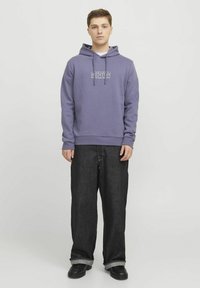 Sudadera morada con logo frontal, cordones y dobladillo acanalado, combinada con jeans negros holgados y calzado negro con suela gruesa.
