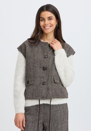 Femme souriante portant un gilet marron boutonné sur un pull en tricot blanc et un pantalon marron assorti, debout devant un fond clair uni.