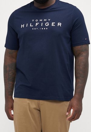 Mężczyzna w granatowym T-shircie Tommy Hilfiger z białym logo i beżowych spodniach, widoczne tatuaże na ramionach.