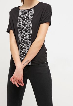 Persoon die een zwart T-shirt met korte mouwen draagt, voorzien van witte geometrische borduursels aan de voorkant, gecombineerd met een zwarte broek en een tatoeage op de pols.