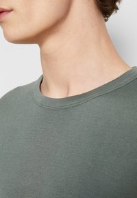 Gros plan sur le cou et la ligne de la mâchoire d'une personne portant un t-shirt gris-vert à col rond, montrant la texture de la peau et le détail du tissu.