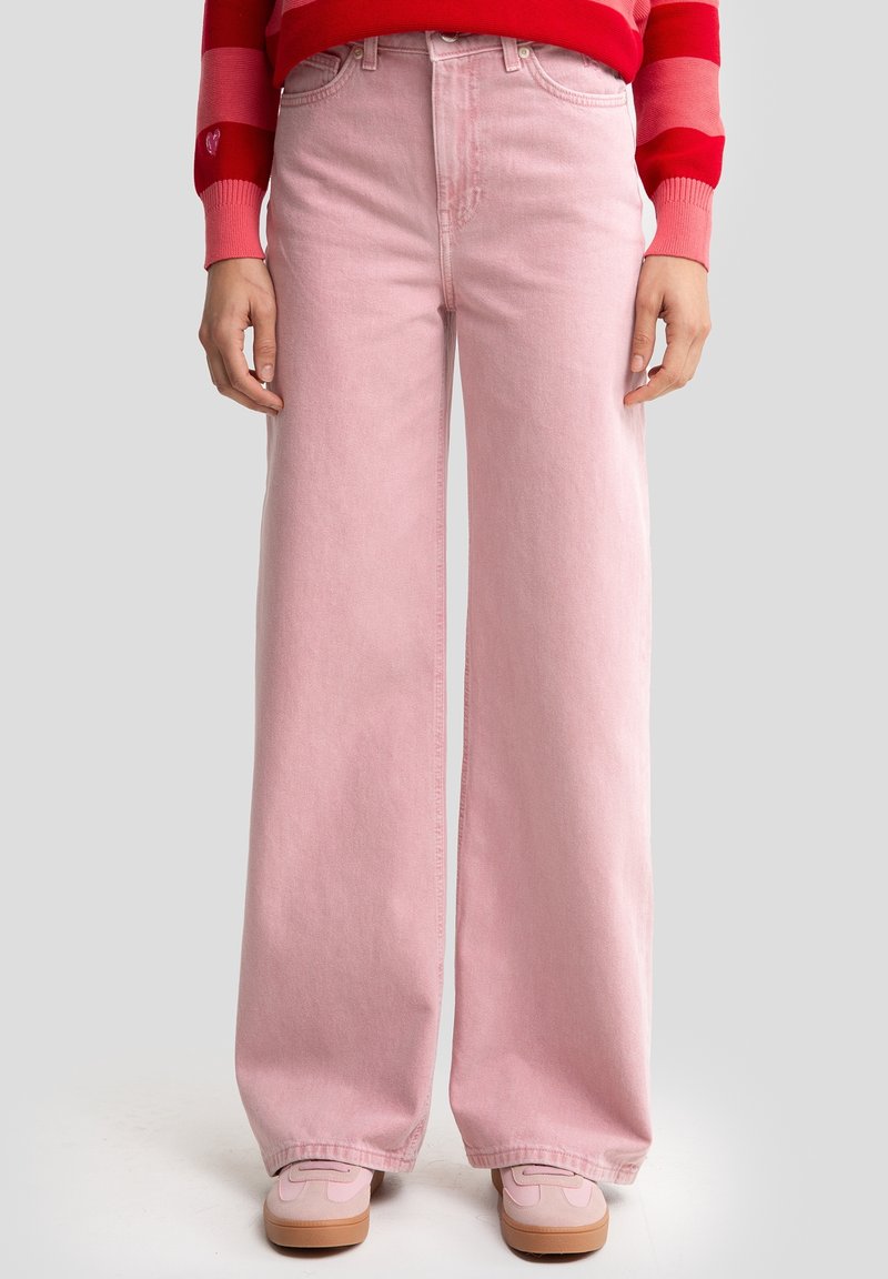 Světle růžové džínové kalhoty s širokými nohavicemi, vysokým pasem a předními kapsami. Kombinované se světle růžovými teniskami.