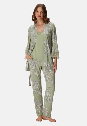 Kvinde stående barfodet iført lysegrønt pyjamasæt med blomsterprint og blondekant på ærmer og halsudskæring, hænder i lommerne.
