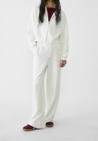 Sudadera blanca con cremallera y diseño corto, combinada con pantalones holgados blancos y zapatillas burdeos. Tejido suave con cinturilla elástica.