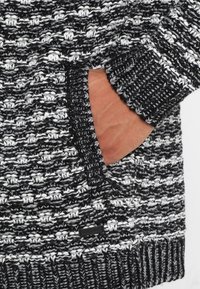 Maglione lavorato a maglia in bianco e nero, con motivo testurizzato. Include una tasca laterale con etichetta accentata nera e polsini a costine.