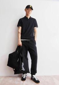 Schwarzes Poloshirt und Jogginghosen, trägt zwei schwarze Einkaufstaschen. Schwarze Sneakers mit weißen Schnürsenkeln. Tätowierungen auf den Armen sichtbar. Einfacher, lässiger Look.