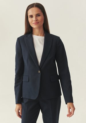 Kvinde med glat brunt hår iført en marineblå blazer og matchende bukser over en hvid top, stående mod en ensartet lys baggrund.