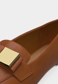 Chaussure plate en cuir marron avec une grande boucle carrée en métal doré sur la partie supérieure avant.