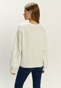 Witte trui met textuur, bloemmotieven op de mouwen. Geribbelde zoom en manchetten. Gecombineerd met blauwe jeans. Schone, minimalistische stijl.