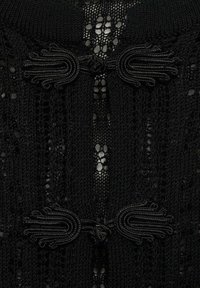 Schwarzes gestricktes Kleidungsstück mit strukturiertem, offenem Gewebe, kunstvollem Rand mit geschlungenen Akzenten und einem auffälligen zentralen Verschlussdetails.
