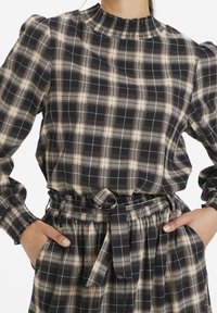 Plaid blouse featuring a smocked neckline, long sleeves, and gathered waist with a belt. Colors include navy, cream, and beige check patterns.