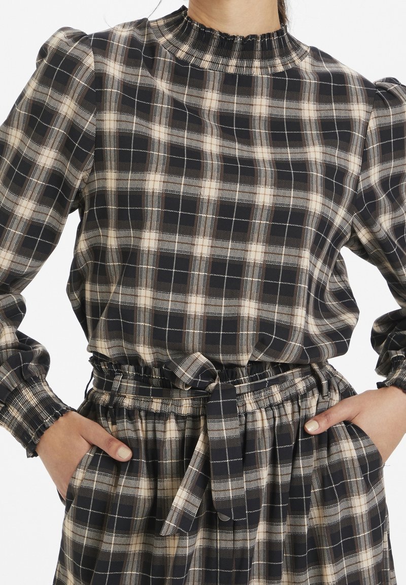 Plaid blouse featuring a smocked neckline, long sleeves, and gathered waist with a belt. Colors include navy, cream, and beige check patterns.