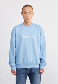 Sudadera azul claro con puños y dobladillo de canalé. Presenta un logo bordado de "carhartt." en el pecho. Se lleva con jeans azules.