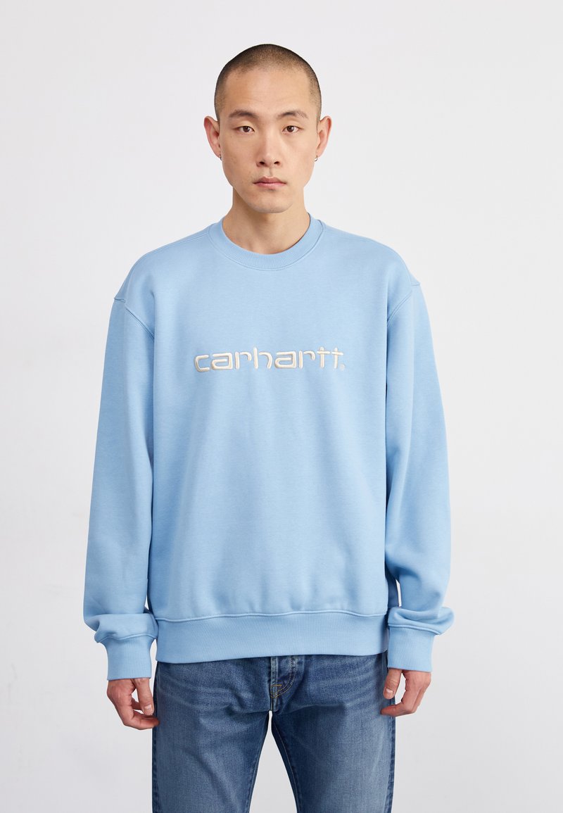 Sudadera azul claro con puños y dobladillo de canalé. Presenta un logo bordado de "carhartt." en el pecho. Se lleva con jeans azules.