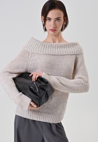 Maglione beige chiaro lavorato a maglia con scollatura larga e a coste che lascia le spalle scoperte, maniche lunghe e vestibilità ampia, abbinato a una borsa nera con texture.