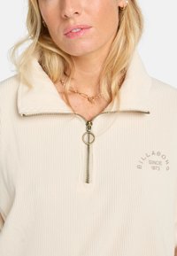 Cremefarbener gerippter Pullover mit einem Reißverschlusskragen, rundem goldenen Reißverschluss und gesticktem "BILLABONG"-Logo. Weicher Stoff und lässige Passform.