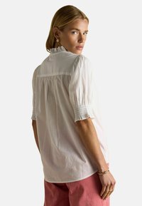 Frau mit blonden Haaren, die eine weiße Bluse mit Puffärmeln und Rüschensaum, pinkfarbene Hosen, goldene Ohrringe und ein Armband trägt und über die Schulter schaut.