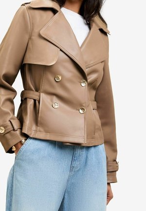 Femme portant une veste en cuir beige croisée à boutons dorés et un jean bleu clair, mains dans les poches, chemise blanche en dessous.