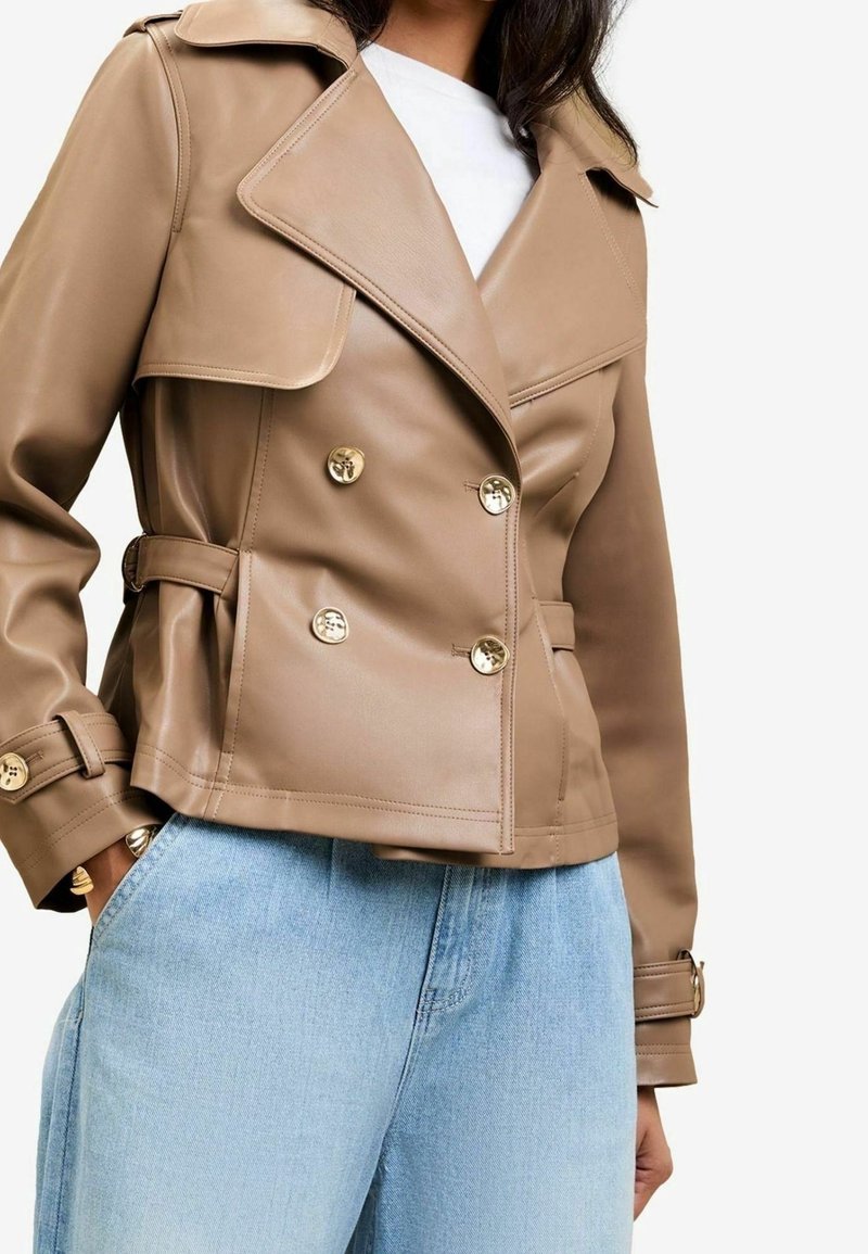 Femme portant une veste en cuir beige croisée à boutons dorés et un jean bleu clair, mains dans les poches, chemise blanche en dessous.