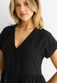 Black dress with a v-neckline and short sleeves, featuring a tiered, gathered skirt for a relaxed silhouette. Smooth, soft fabric texture.